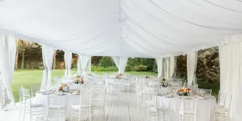 A large white event tent with draped fabric, set up with round tables and chairs for a wedding or party.