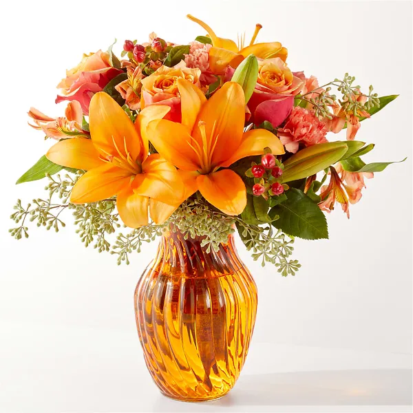A floral arrangement featuring orange lilies, roses, and greenery in an amber glass vase.