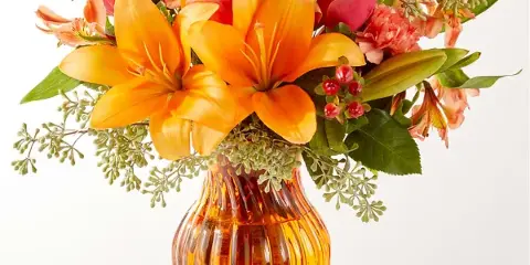 A floral arrangement featuring orange lilies, roses, and greenery in an amber glass vase.