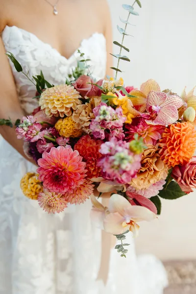 A bride in a white lace wedding dress holding a large, colorful bouquet of dahlias, roses, and orchids.