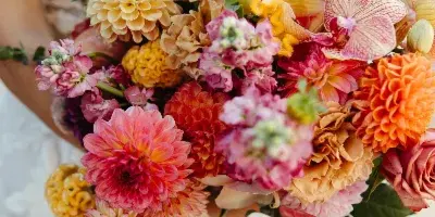 A bride in a white lace wedding dress holding a large, colorful bouquet of dahlias, roses, and orchids.