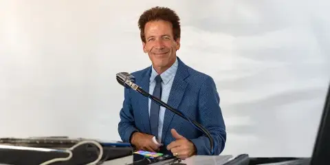 A professional DJ in a blue suit standing behind a mixing console and microphone.