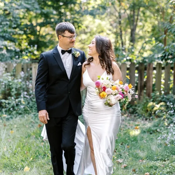 A bride in a white gown holding a vibrant, garden-inspired bouquet with pink, yellow, and white flowers while walking with her groom in a green outdoor setting.