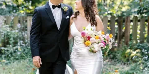 A bride in a white gown holding a vibrant, garden-inspired bouquet with pink, yellow, and white flowers while walking with her groom in a green outdoor setting.