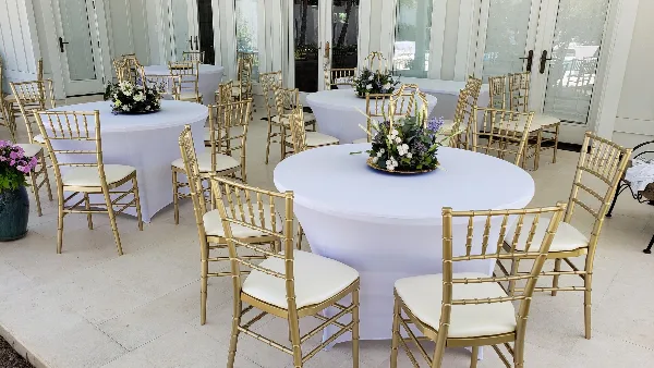 An outdoor event space set up with round tables covered in white tablecloths and surrounded by gold Chiavari chairs.