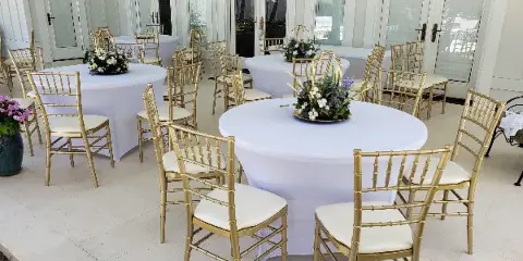 An outdoor event space set up with round tables covered in white tablecloths and surrounded by gold Chiavari chairs.