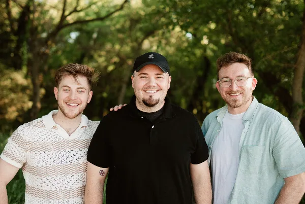 Three smiling men standing together outdoors in front of trees, representing the River City Visuals team.