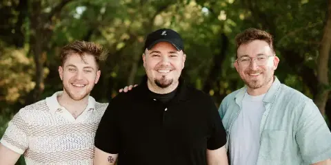 Three smiling men standing together outdoors in front of trees, representing the River City Visuals team.