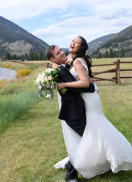 A bride and groom laughing and embracing in a grassy field with mountains in the background.