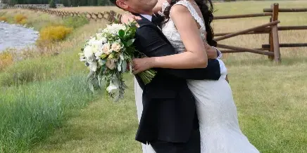 A bride and groom laughing and embracing in a grassy field with mountains in the background.