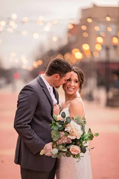 A bride and groom embracing outdoors with string lights in the background, the bride holding a floral bouquet.
