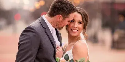 A bride and groom embracing outdoors with string lights in the background, the bride holding a floral bouquet.