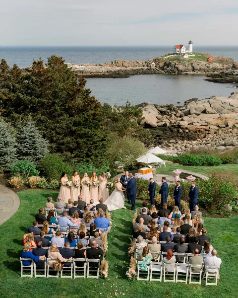Aerial view of a wedding ceremony on a grassy lawn overlooking the ocean and a lighthouse, with the bride and groom at the altar and guests seated in rows.