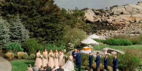 Aerial view of a wedding ceremony on a grassy lawn overlooking the ocean and a lighthouse, with the bride and groom at the altar and guests seated in rows.