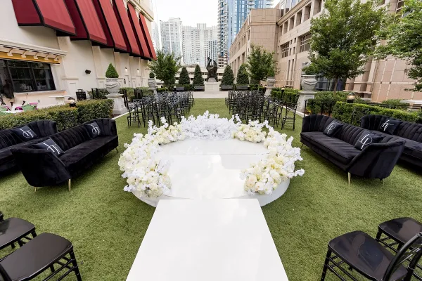 Outdoor event setup with black velvet lounge sofas, black chiavari chairs, a round white dance floor ringed by white flowers, and a white aisle runner on artificial turf.