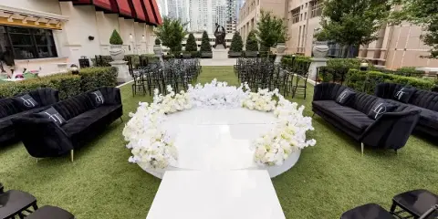 Outdoor event setup with black velvet lounge sofas, black chiavari chairs, a round white dance floor ringed by white flowers, and a white aisle runner on artificial turf.