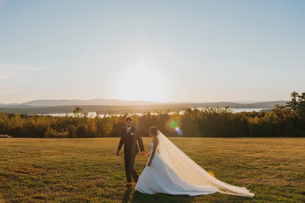 A bride and groom walking hand-in-hand across a grassy field at sunset with a lake and mountains in the background.