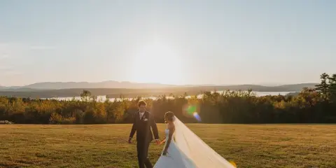 A bride and groom walking hand-in-hand across a grassy field at sunset with a lake and mountains in the background.