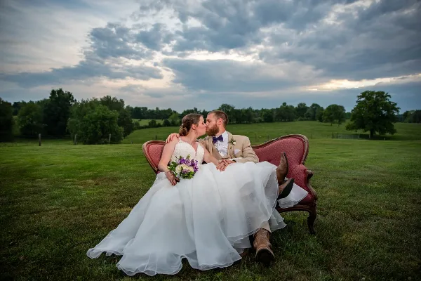 A bride and groom kissing while seated on a red vintage couch in a grassy field under a cloudy sky.