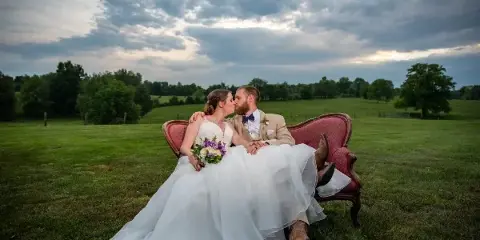 A bride and groom kissing while seated on a red vintage couch in a grassy field under a cloudy sky.
