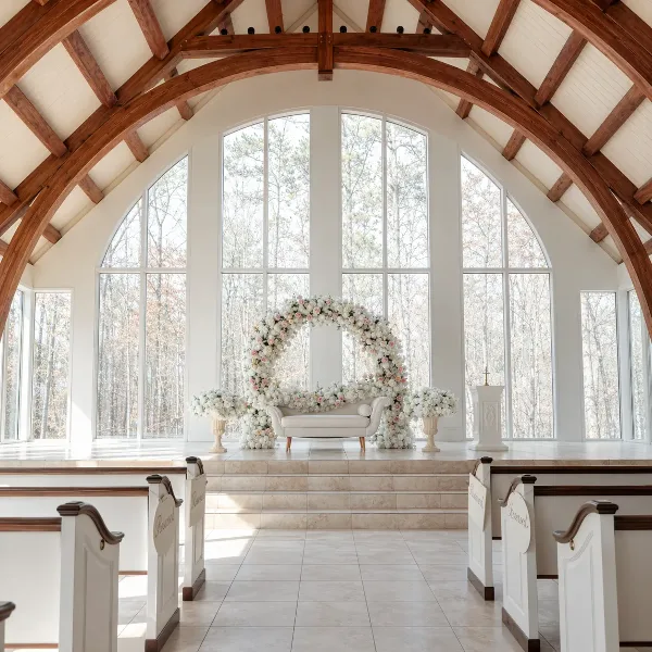 A floral arch and white sofa arranged on a stage in a chapel with large arched windows.