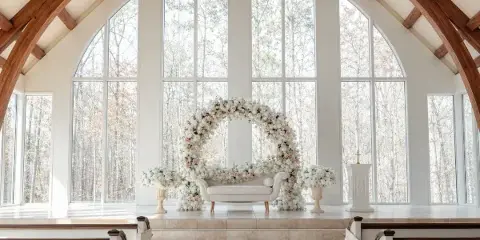 A floral arch and white sofa arranged on a stage in a chapel with large arched windows.