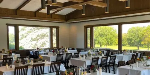 An indoor wedding banquet hall with white tablecloths and large windows overlooking a green landscape.