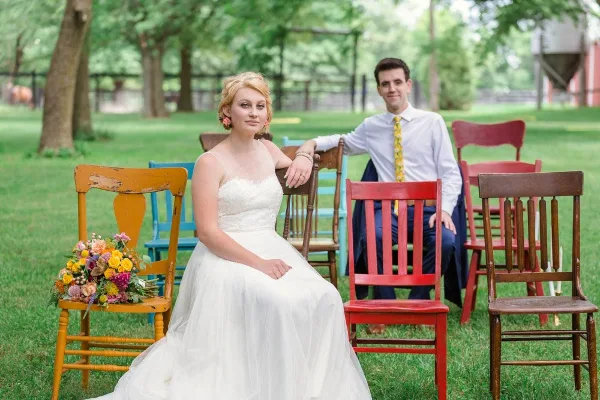 A bride and groom posing outdoors among colorful wooden chairs with a bouquet of flowers.
