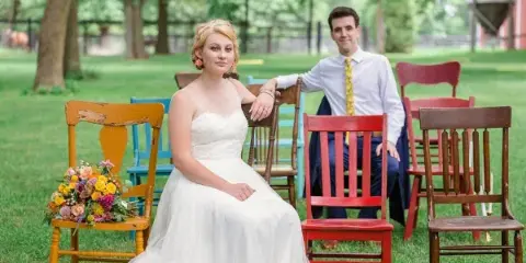 A bride and groom posing outdoors among colorful wooden chairs with a bouquet of flowers.