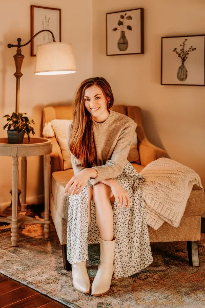 A woman seated on a sofa in a warmly lit room with framed botanical prints, a floor lamp, and a patterned rug.