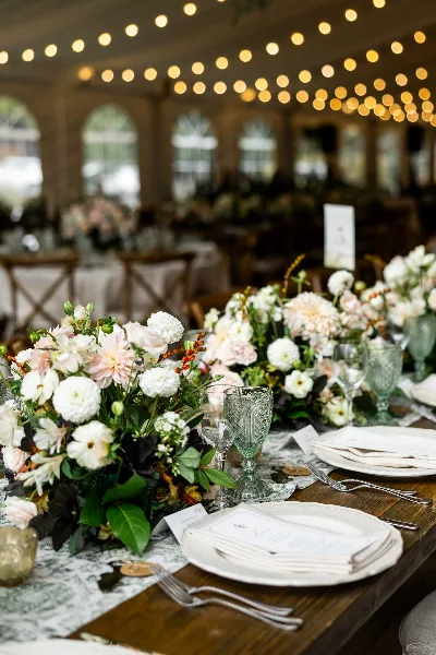 A wedding table setting featuring lush floral centerpieces in white and blush, paired with textured green glassware and white plates under warm string lights.