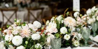 A wedding table setting featuring lush floral centerpieces in white and blush, paired with textured green glassware and white plates under warm string lights.