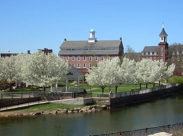 Exterior view of the historic brick Belknap Mill building with a cupola, surrounded by blooming trees and a river.