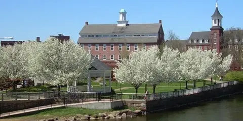 Exterior view of the historic brick Belknap Mill building with a cupola, surrounded by blooming trees and a river.