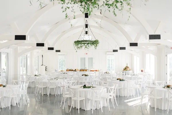 A white event hall set up with numerous round tables covered in white linens, surrounded by white cross-back chairs and a buffet table in the background.