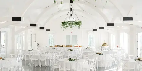 A white event hall set up with numerous round tables covered in white linens, surrounded by white cross-back chairs and a buffet table in the background.