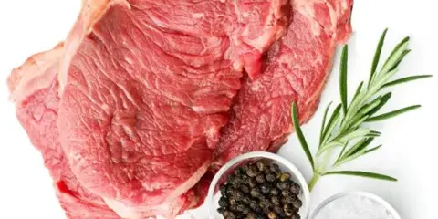A large raw cut of red meat displayed with fresh rosemary, garlic, salt, and peppercorns.