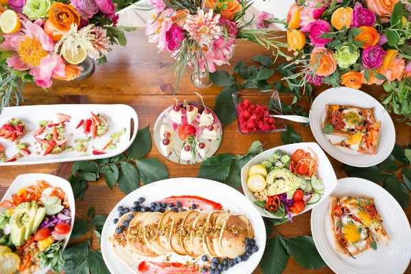 A top-down view of a catering table set with colorful floral arrangements, fresh fruit tarts, a large platter of pancakes with berries, and various vegetable salads.