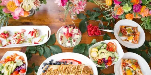 A top-down view of a catering table set with colorful floral arrangements, fresh fruit tarts, a large platter of pancakes with berries, and various vegetable salads.