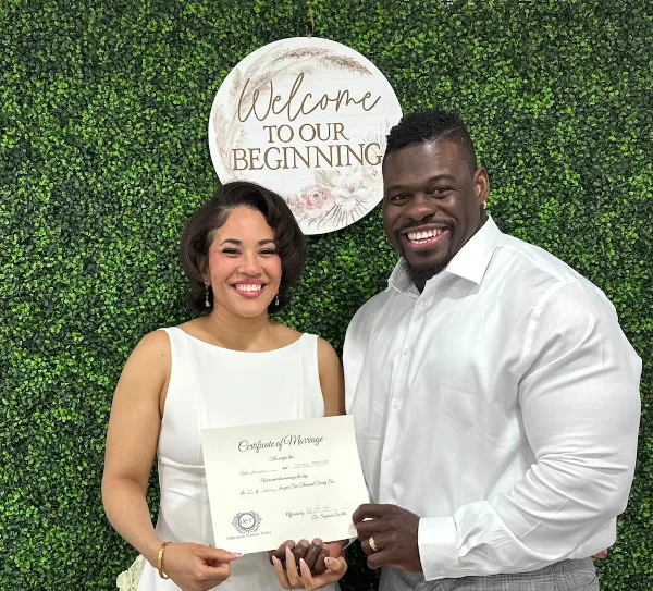 A smiling couple in white attire holding a marriage certificate with the Officiantly Forever Yours seal in front of a greenery wall and a 'Welcome to Our Beginning' sign.