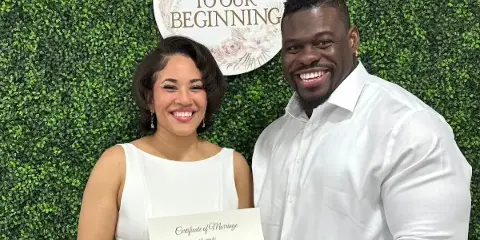 A smiling couple in white attire holding a marriage certificate with the Officiantly Forever Yours seal in front of a greenery wall and a 'Welcome to Our Beginning' sign.