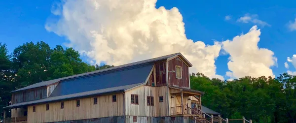 Exterior view of a large wooden barn with a metal roof set against a blue sky and trees.