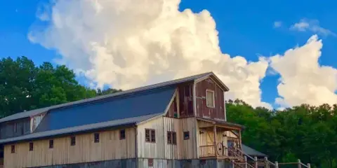 Exterior view of a large wooden barn with a metal roof set against a blue sky and trees.