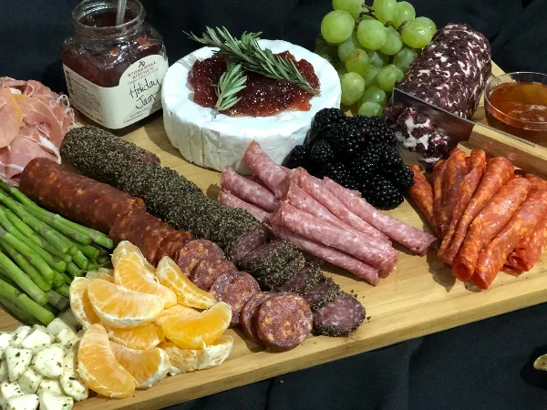 A catering spread featuring a charcuterie board with various cured meats, a wheel of cheese with jam, fresh fruit, and asparagus.