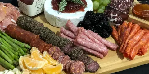 A catering spread featuring a charcuterie board with various cured meats, a wheel of cheese with jam, fresh fruit, and asparagus.