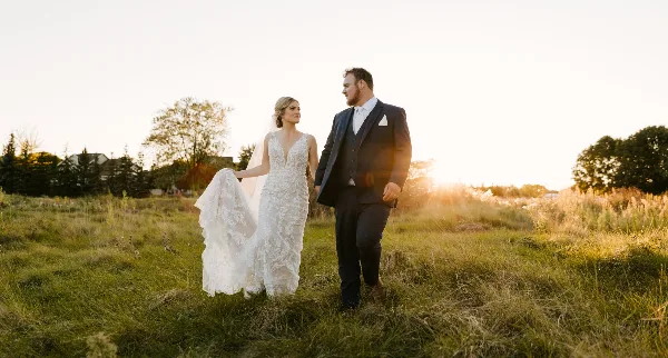 A bride and groom walking through a grassy field at sunset.