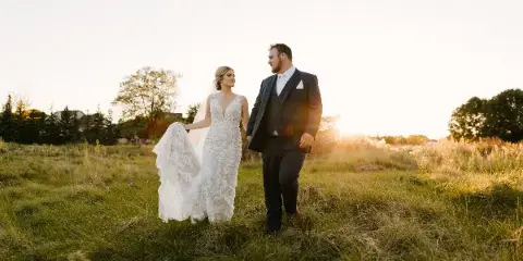 A bride and groom walking through a grassy field at sunset.
