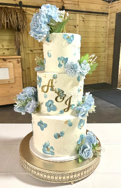 A three-tier white wedding cake decorated with blue floral accents and gold monogram initials, displayed on a gold stand.
