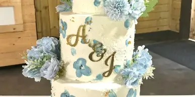 A three-tier white wedding cake decorated with blue floral accents and gold monogram initials, displayed on a gold stand.