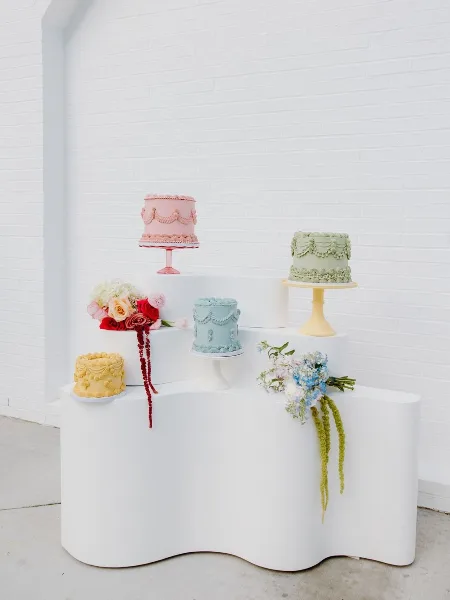A display of various custom cakes and pastries arranged on a white tiered stand against a white brick wall.